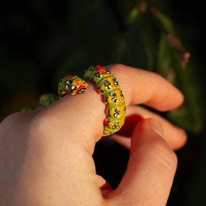 May include: A quirky caterpillar ring, with a yellow and green segmented body, and black and red detailing. The ring is worn on a finger, highlighting its unusual design. The ring's design is inspired by nature and insects.