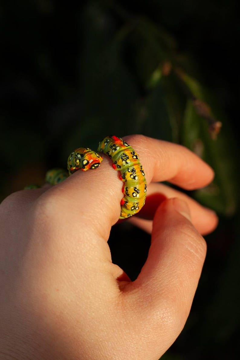 May include: A quirky caterpillar ring, with a yellow and green segmented body, and black and red detailing. The ring is worn on a finger, highlighting its unusual design. The ring's design is inspired by nature and insects.