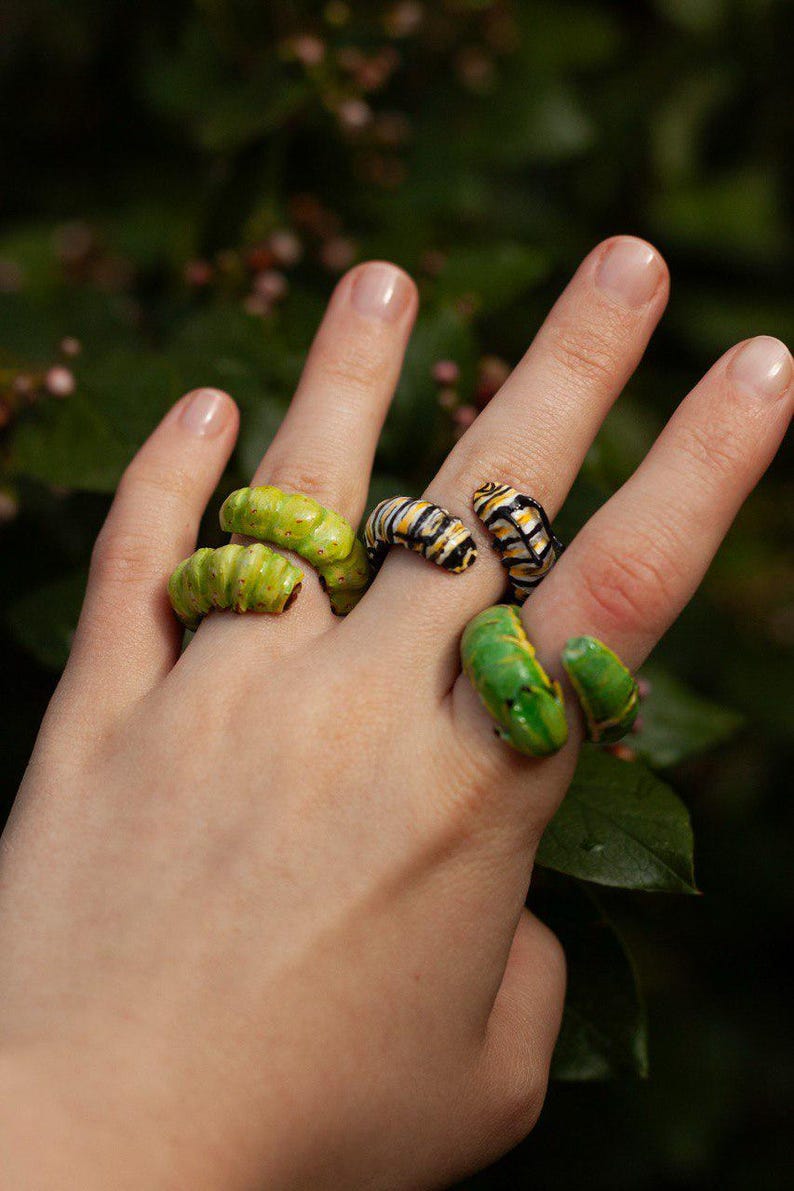 May include: A hand adorned with several rings shaped like caterpillars. The rings are in shades of green, yellow, black, and white. The rings appear to be made from a painted or glazed material, showcasing intricate details.