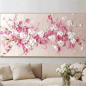 Puede incluir: Una gran pintura abstracta de flores rosas y blancas sobre un fondo rosa claro. La pintura está enmarcada en un marco dorado y está colgada en una pared sobre un sofá blanco.