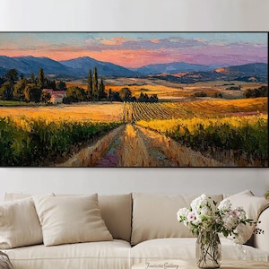 Könnte beinhalten: Ölgemälde einer toskanischen Landschaft mit einer Straße durch goldene Felder zu einem Dorf und Bergen unter einem Sonnenuntergangshimmel. Das Kunstwerk ist schwarz gerahmt und hängt über einem beigen Sofa.