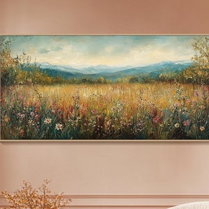 May include: A painting of a field of wildflowers with a mountain range in the background. The painting is in a gold frame and is hanging on a pink wall.