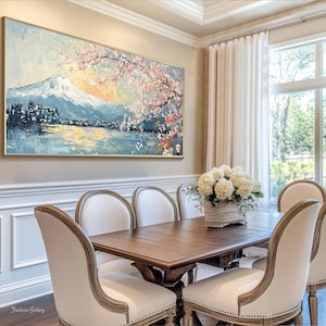 May include: A dining room with a large painting of a mountain scene with cherry blossoms above a lake. The painting is framed in gold and hangs on a beige wall. The room has a wooden table with six chairs, a vase of white flowers, and a large window with white curtains.