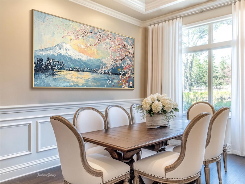 May include: A dining room with a large painting of a mountain scene with cherry blossoms above a lake. The painting is framed in gold and hangs on a beige wall. The room has a wooden table with six chairs, a vase of white flowers, and a large window with white curtains.
