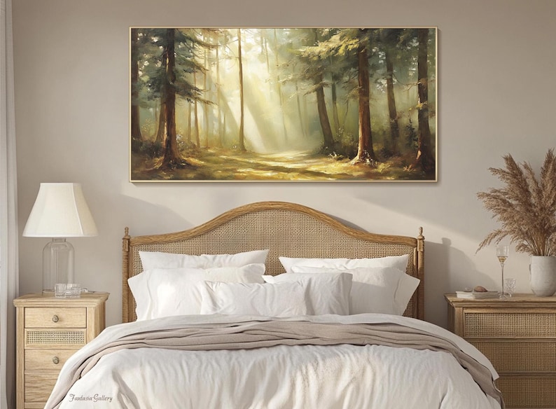 May include: A painting of a sunlit forest scene with tall trees and a path leading into the woods. The painting is framed in a gold frame and hangs above a bed with white linens.