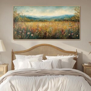 May include: A painting of a field of wildflowers with a mountain range in the background. The painting is framed and hanging on a wall above a bed with white linens.
