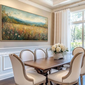 May include: A large landscape painting of a field of wildflowers with mountains in the background hangs on a wall in a dining room. The painting is framed in gold and has a light blue sky. The dining room has a wooden table with six white chairs and a vase of white flowers in the center.