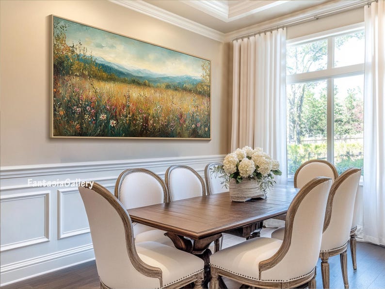 May include: A large landscape painting of a field of wildflowers with mountains in the background hangs on a wall in a dining room. The painting is framed in gold and has a light blue sky. The dining room has a wooden table with six white chairs and a vase of white flowers in the center.