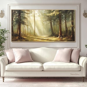 May include: A painting of a sunlit forest scene with tall trees and a path leading into the woods. The painting is framed in a gold frame and hangs on a white wall above a white sofa with pink pillows.