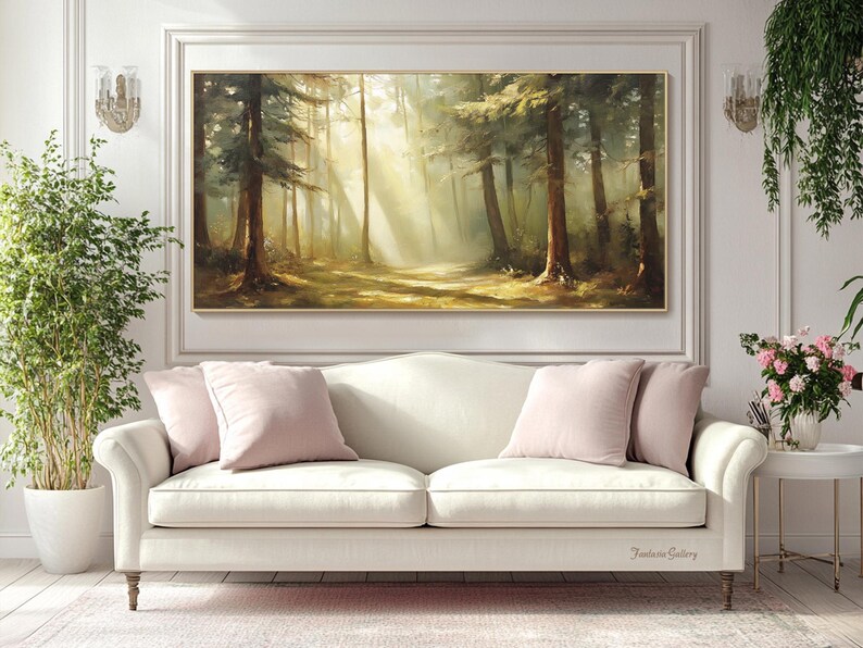 May include: A painting of a sunlit forest scene with tall trees and a path leading into the woods. The painting is framed in a gold frame and hangs on a white wall above a white sofa with pink pillows.