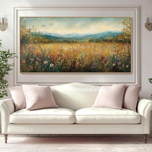 May include: A white sofa with two pink throw pillows in front of a painting of a field of wildflowers and a mountain range in the background.