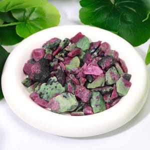 Könnte beinhalten: Eine Ansammlung von rohen, unpolierten Rubin- und Zoisit-Mineralfragmenten mit einer Mischung aus tiefem Magenta, leuchtendem Grün und schwarzen Einschlüssen. Diese kleinen, unregelmäßigen Steinstücke befinden sich in einer runden weißen Schale.