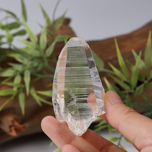 Könnte beinhalten: Ein klares, spitzes Quarzkristall-Exemplar mit ausgeprägten horizontalen Streifen auf der Oberfläche. Das Mineral hat ein durchscheinendes, glasartiges Aussehen mit einer gezackten Basis und einer natürlich geformten Spitze.