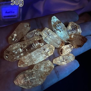 Könnte beinhalten: Eine Ansammlung klarer Quarz-Kristalle, einige mit bernsteinfarbenen Tönen, wird gezeigt. Die Kristalle variieren in Größe und Form, mit sichtbaren inneren Strukturen. Der Text "50g" ist enthalten.