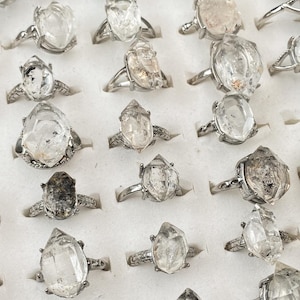 May include: A display of silver rings, each featuring a unique, clear crystal stone. The rings are presented in a white tray, highlighting the variety of crystal shapes and sizes. The silver bands have different designs.