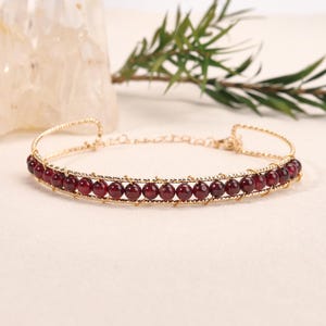 May include: A gold-toned wire bracelet featuring a single row of small, round, deep red garnet-coloured beads. The metal wire is textured and wraps around each bead to secure them in place, with a delicate chain extension at the back closure.