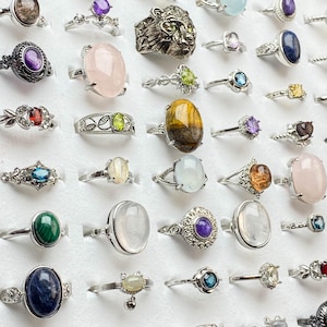 Könnte beinhalten: Eine Sammlung von Silberringen mit verschiedenen Edelsteinen, darunter Amethyst, Rosenquarz und blauer Topas. Die Ringe werden in einem weißen Tablett präsentiert und zeigen eine Vielzahl von Formen und Größen, von zarten Bändern bis hin zu Statement-Stücken.