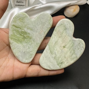 Könnte beinhalten: Zwei herzförmige Gua-Sha-Werkzeuge aus hellgrünem Jade. Die Werkzeuge werden in einer Hand gehalten, mit einem weichen, weißen Stoff und einer kleinen Sukkulentenpflanze im Hintergrund. Die Werkzeuge sind etwa 10 cm lang.