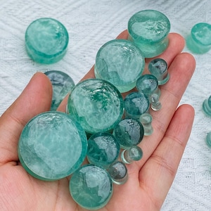 Può includere: Una collezione di tappi e tunnel in pietra traslucida color verde acqua di varie dimensioni. I tappi sono rotondi e lisci, con una leggera curvatura. I tunnel sono anch'essi rotondi, con un centro cavo. Gli oggetti sono tenuti in una mano.
