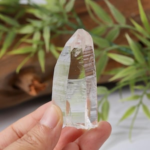 Könnte beinhalten: Ein klarer, spitzer Quarzkristall mit natürlichen horizontalen Streifen entlang seiner vertikalen Facetten. Das Mineral zeigt ein transparentes, glasartiges Aussehen mit sichtbaren inneren Einschlüssen und einer scharfen, natürlich geformten Spitze.