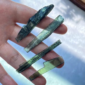 Pode incluir: Quatro espécimes de cristais de cianita brutos e alongados com uma coloração translúcida de verde profundo a azul-esverdeado. Os cristais exibem uma estrutura distinta em lâminas com estrias naturais e graus variados de transparência.