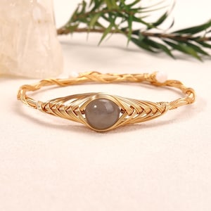 May include: A gold-toned wire-wrapped bracelet featuring a central round, smooth, grey stone. The metal wire is intricately coiled and woven around the stone and continues to form the circular band, which is finished with small white beads.