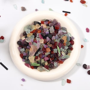 Könnte beinhalten: Eine runde, cremeweiße Keramikschale gefüllt mit einer Sammlung kleiner, roher, mehrfarbiger Edelsteine und Mineralfragmente in Rot-, Lila-, Schwarz-, Hellgrün- und durchscheinenden Orangetönen, mit verstreuten losen Steinen um die Schale.