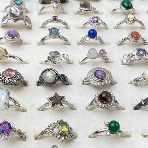 Op de afbeelding: Een verzameling diverse metalen ringen gerangschikt in een raster, met verschillende ontwerpen waaronder cabochon edelstenen, bloemmotieven, stervormen en filigraan banden met kleurrijke stenen in blauwe, groene, paarse en heldere tinten.