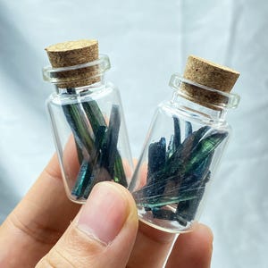 Puede incluir: Dos pequeños frascos de vidrio transparente con tapones de corcho, cada uno lleno de fragmentos de cristal azul verdoso oscuro. Los frascos se sostienen en una mano sobre un fondo blanco suave. Los cristales tienen una calidad iridiscente brillante.