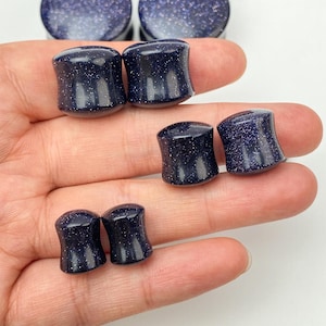 Könnte beinhalten: Eine Sammlung dunkelblauer Ohrstöpsel mit einem gesprenkelten, glitzernden Aussehen. Die Stöpsel sind zylindrisch mit ausgestellten Kanten. Das Bild zeigt mehrere Paare von Stöpseln in verschiedenen Größen, mit einigen größeren, runden Stöpseln im Hintergrund.