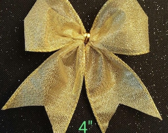 May include: A gold-colored decorative bow with a shimmering, textured appearance. The bow is approximately 4 inches in width, with a central knot and pointed ends. The background is black with small white specks.