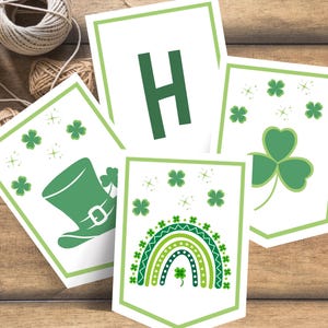 May include: A set of St. Patrick's Day banner cards. Each card has a white background with green borders and green graphics, such as a leprechaun hat, shamrocks, a rainbow, and the letter "H". The cards are displayed on a wooden surface.