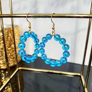 Peut inclure: Une paire de boucles d'oreilles créoles en perles bleues avec des crochets dorés. Les perles sont irisées et de couleur bleu clair.