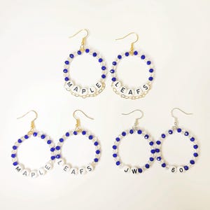 Può includere: Una collezione di orecchini a cerchio. Ogni orecchino presenta un design circolare con perline blu e perline con lettere bianche che compongono "MAPLE LEAFS" e iniziali. Gli orecchini hanno ganci color oro o argento.
