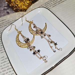 Puede incluir: Pendientes de media luna dorados con cadenas colgantes y abalorios de estrellas. Las cadenas están adornadas con diamantes de imitación transparentes.