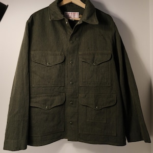 Puede incluir: Chaqueta verde oscuro con cuatro bolsillos delanteros y botones. La chaqueta tiene cuello y est&aacute; colgada de una percha de madera. La chaqueta est&aacute; hecha de una tela texturizada y parece ser una prenda de abrigo duradera.