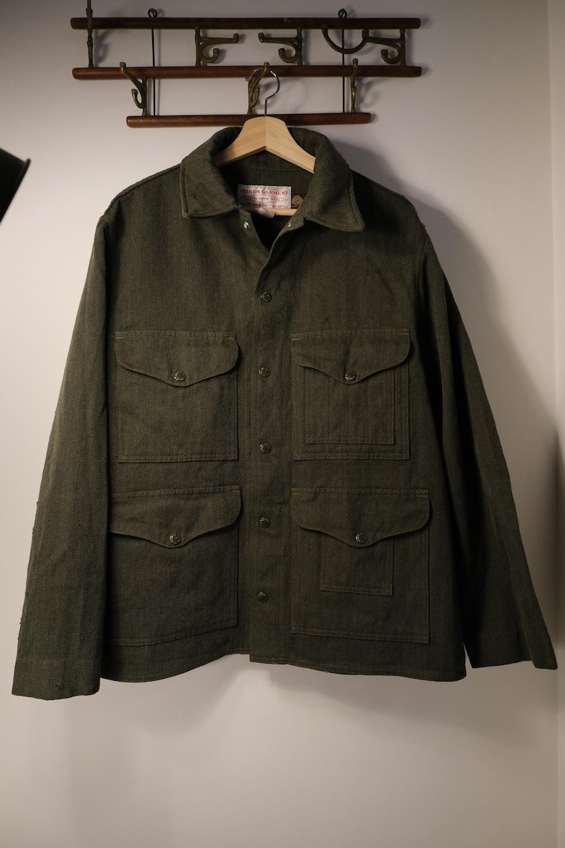 Puede incluir: Chaqueta verde oscuro con cuatro bolsillos delanteros y botones. La chaqueta tiene cuello y est&aacute; colgada de una percha de madera. La chaqueta est&aacute; hecha de una tela texturizada y parece ser una prenda de abrigo duradera.