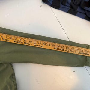 May include: An olive green long-sleeved shirt with a wooden ruler laid across the sleeve. The ruler displays the text "Window Shades, Venetian Blinds, Draperies" and "WILLIAM VOLKER & COMPANY". The shirt's sleeve measures approximately 25 inches.