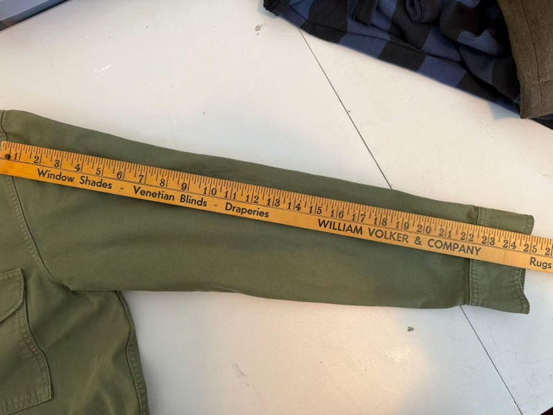 May include: An olive green long-sleeved shirt with a wooden ruler laid across the sleeve. The ruler displays the text "Window Shades, Venetian Blinds, Draperies" and "WILLIAM VOLKER & COMPANY". The shirt's sleeve measures approximately 25 inches.
