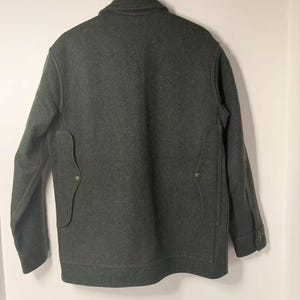 May include: Dark green wool jacket with a button-down front, two front pockets, and buttoned cuffs. The jacket is hanging on a wooden hanger against a white wall. The jacket's back is visible.