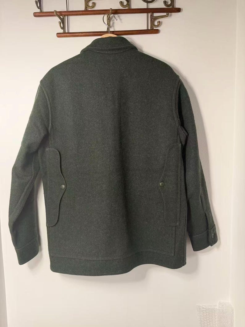 May include: Dark green wool jacket with a button-down front, two front pockets, and buttoned cuffs. The jacket is hanging on a wooden hanger against a white wall. The jacket's back is visible.