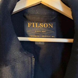 Puede incluir: Primer plano de una chaqueta azul marino colgada de una percha de madera. La chaqueta tiene cierre de cremallera y cuello. La etiqueta dice "FILSON SINCE 1897 C.C. FILSON CO. - SEATTLE, WA". Talla M.