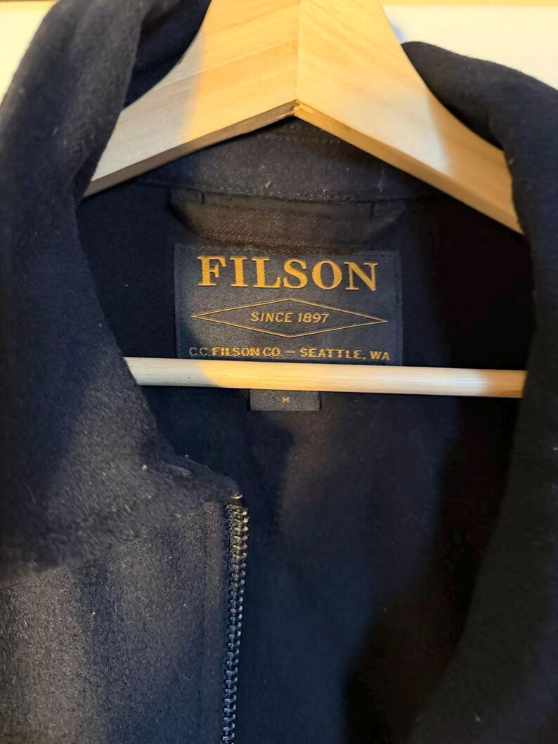 Puede incluir: Primer plano de una chaqueta azul marino colgada de una percha de madera. La chaqueta tiene cierre de cremallera y cuello. La etiqueta dice "FILSON SINCE 1897 C.C. FILSON CO. - SEATTLE, WA". Talla M.
