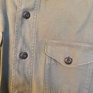 May include: Close-up of an olive green button-down shirt. The shirt features a buttoned front, a chest pocket, and brown buttons. The fabric has visible stitching details.