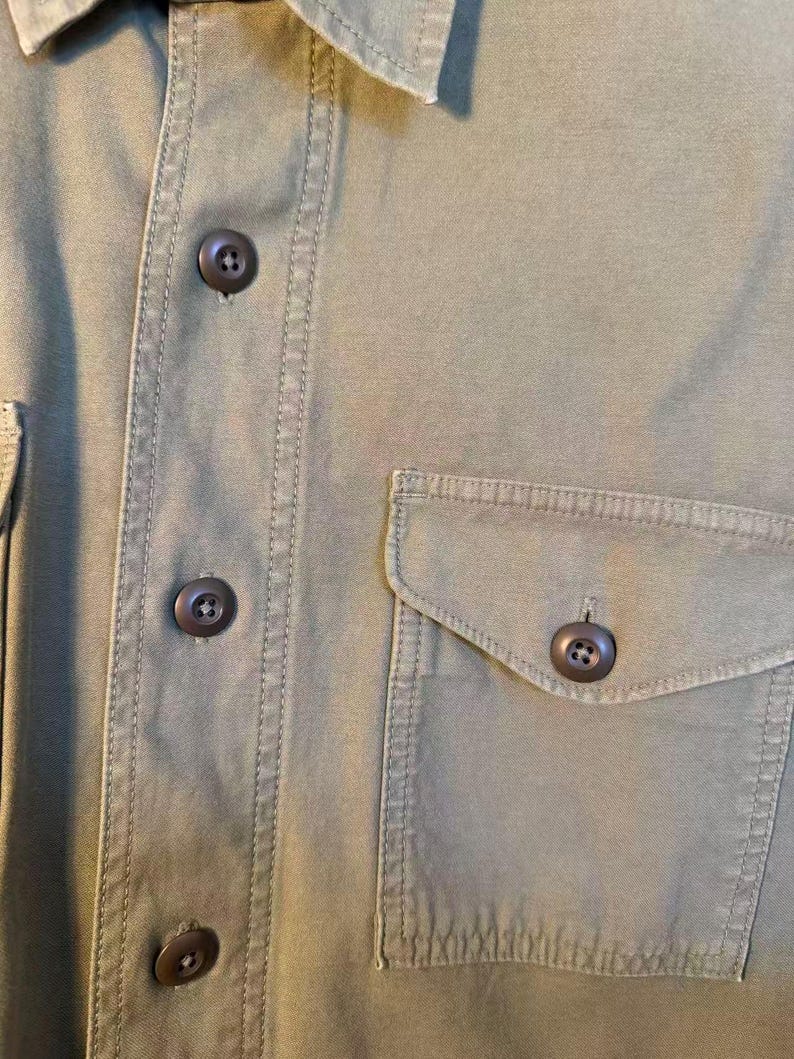May include: Close-up of an olive green button-down shirt. The shirt features a buttoned front, a chest pocket, and brown buttons. The fabric has visible stitching details.
