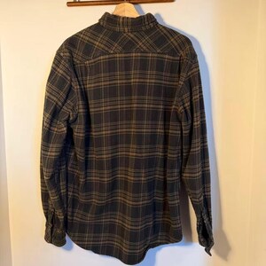 May include: A long-sleeved, button-down flannel shirt in a black and brown plaid pattern. The shirt is hanging on a wooden hanger, showcasing the back of the garment. The fabric appears to be soft and comfortable.
