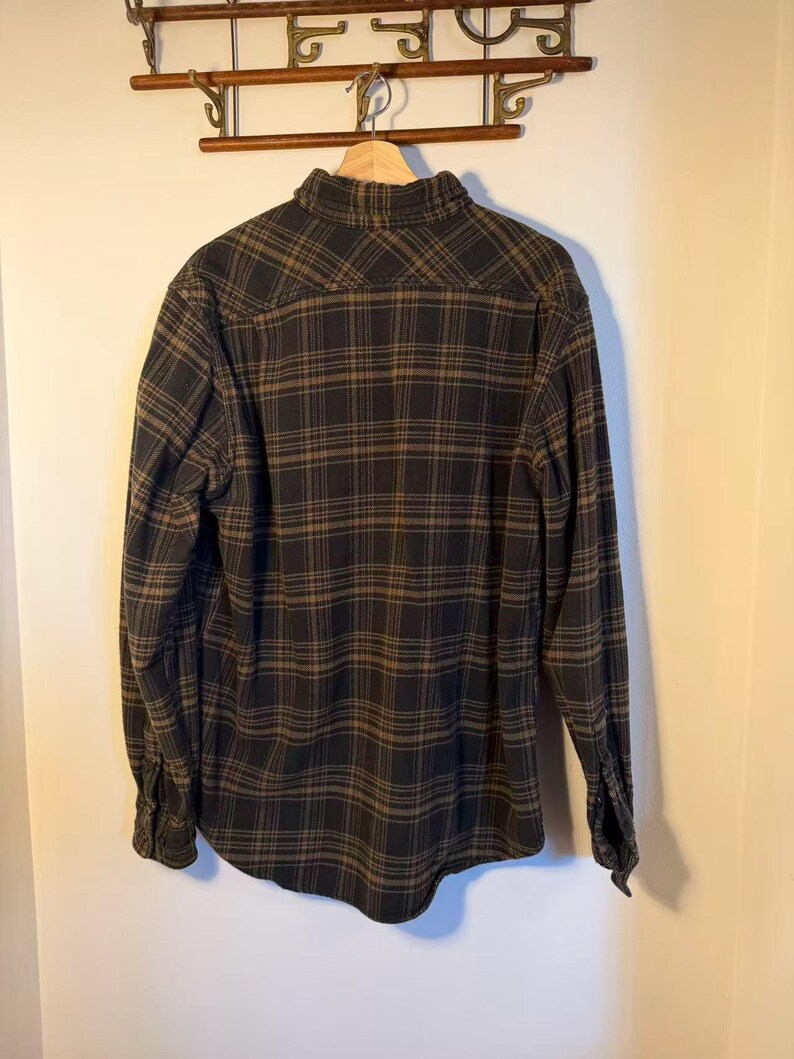 May include: A long-sleeved, button-down flannel shirt in a black and brown plaid pattern. The shirt is hanging on a wooden hanger, showcasing the back of the garment. The fabric appears to be soft and comfortable.