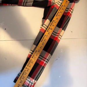 以下が含まれることがあります： 赤、黒、クリーム、グレーのチェック柄長袖シャツ。 シャツはクラシックなボタンダウンデザインです。 木製の定規にテキストが書かれており、シャツの上に置かれています。 定規には「Window Shades - Venetian Blinds - Draperies」と「WILLIAM VOLKER & COMPANY」と書かれています。