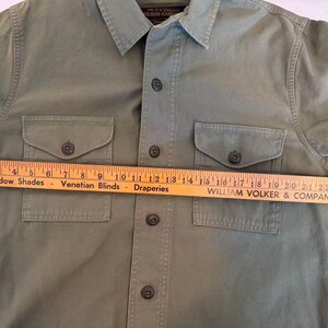 May include: Olive green button-up shirt with two chest pockets and a collared neckline. A wooden ruler, marked in inches, is laid across the shirt. The ruler reads "Window Shades Venetian Blinds Draperies WILLIAM VOLKER & COMPANY".