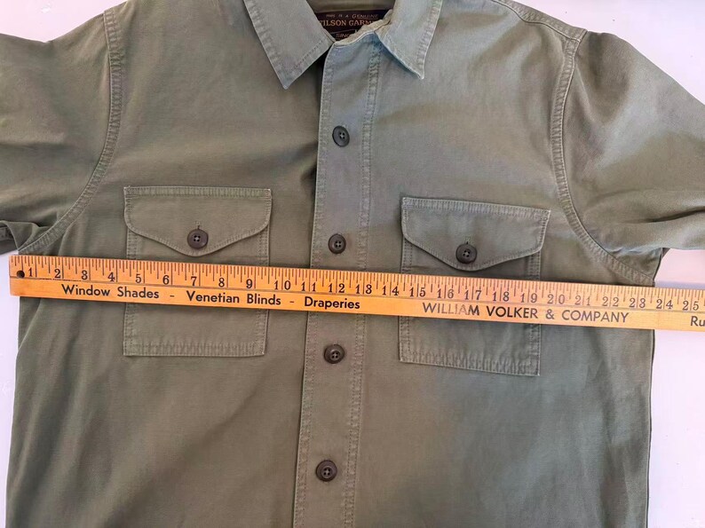 May include: Olive green button-up shirt with two chest pockets and a collared neckline. A wooden ruler, marked in inches, is laid across the shirt. The ruler reads "Window Shades Venetian Blinds Draperies WILLIAM VOLKER & COMPANY".